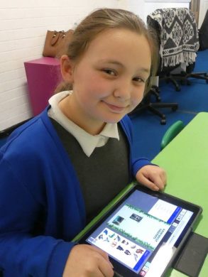 A child smiles as she writes a geography fact file on ainforest Habitats on a tablet using Purple Mash