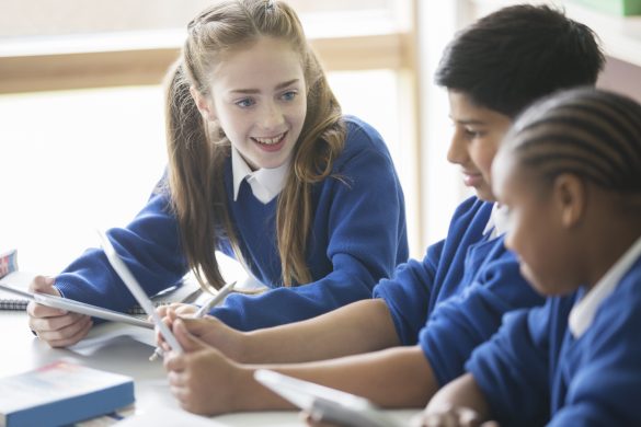 Three students sitting at a desk, one on their tablet.